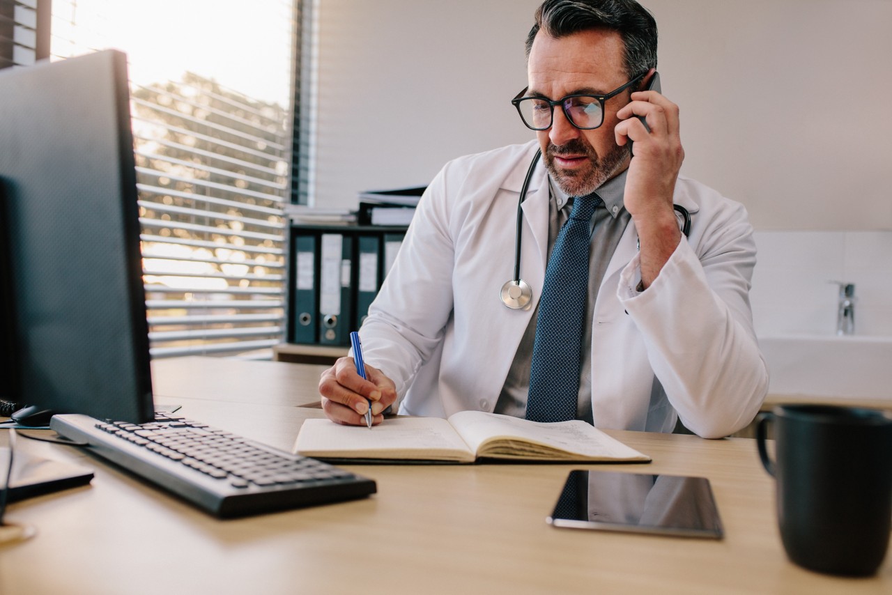 Doctor talking over his mobile phone and making notes in a book. Medicine professional talking on phone and writing in his diary.